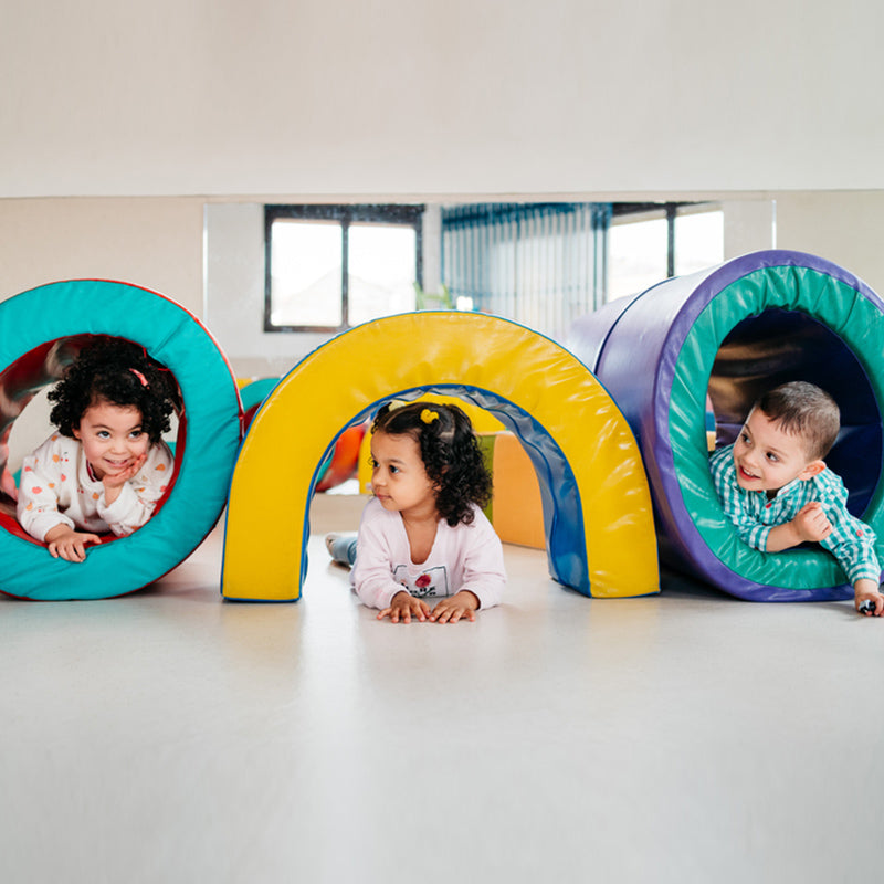 Three young children crawling through colorful foam tunnels in a bright indoor play area