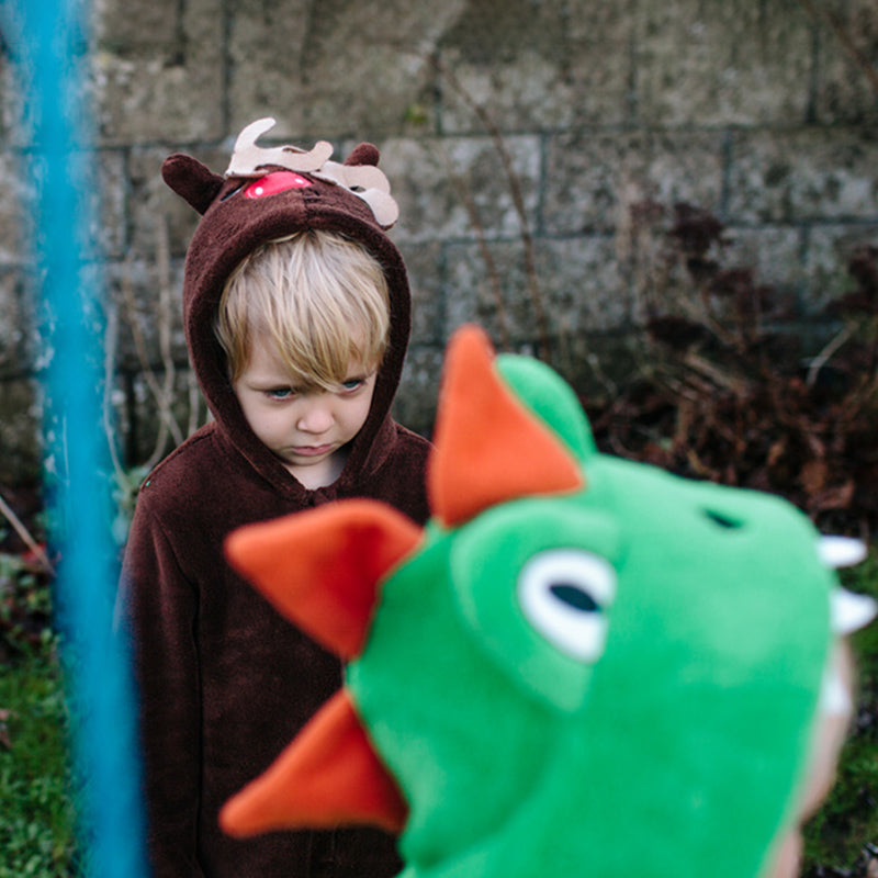 Upset child in brown reindeer hoodie with blurred child in a green dinosaur costume in a backyard
