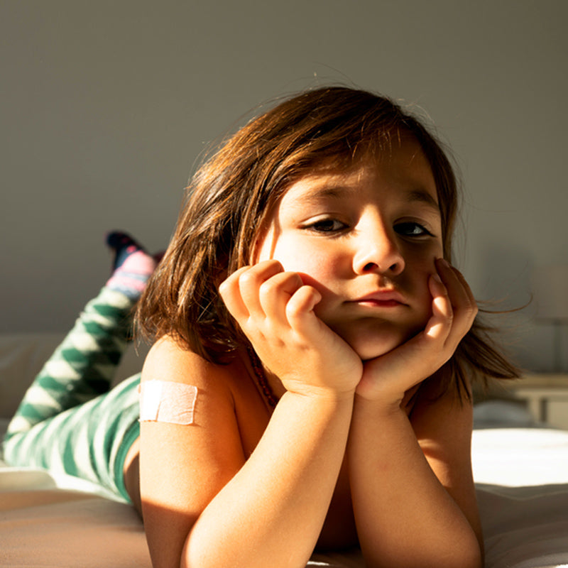 Child lying on a bed propped on hands, looking bored in a sunlit bedroom