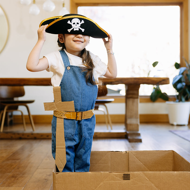 Child in denim overalls wearing a pirate hat and cardboard sword standing in a cardboard box in a sunlit dining room