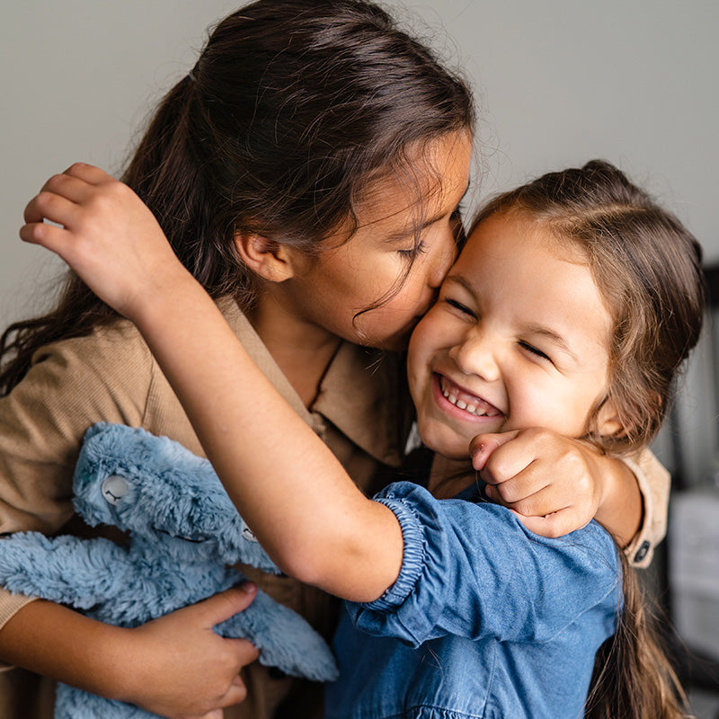 Older sister hugging and kissing her younger sister as the younger smiles and holds a blue plush rabbit