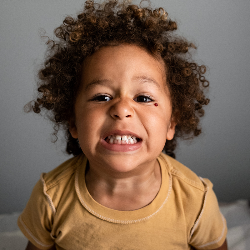 Young child with curly hair scowling and baring teeth toward the camera against a gray background