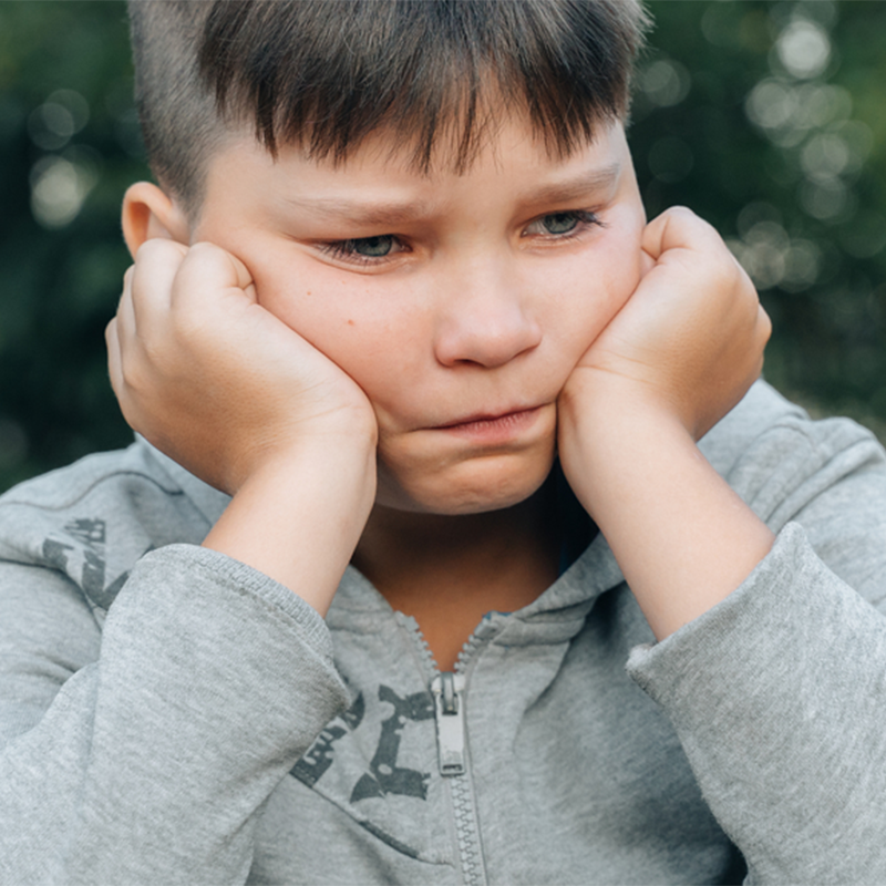 Sad child holding his cheeks in a gray hoodie against a blurred outdoor background