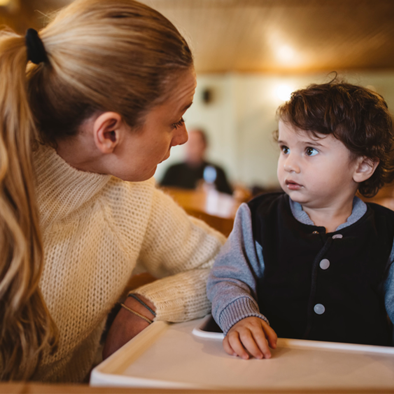 Caregiver leaning toward a toddler in a high chair, comforting him at a warm-lit dining table