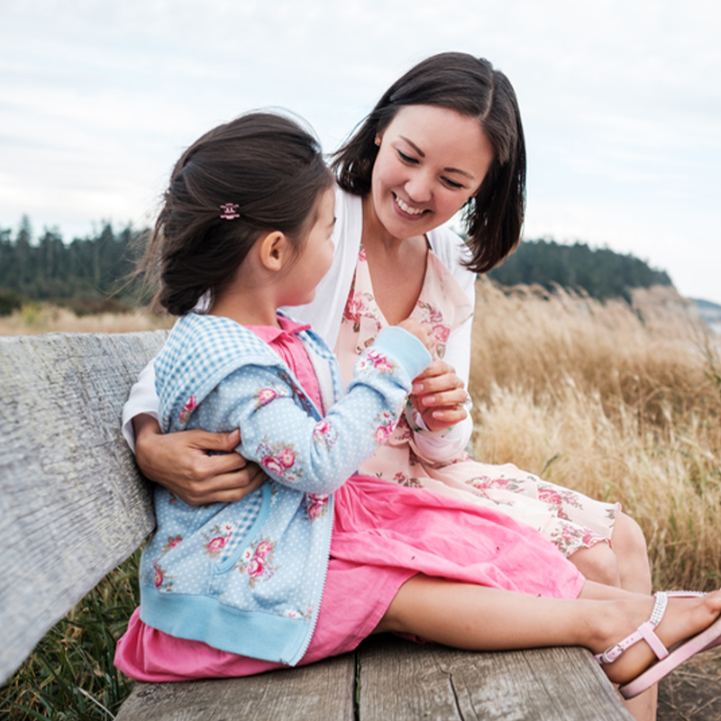 Mother comforting and smiling at her young daughter while they sit on a wooden bench by the beach with tall grass in the background