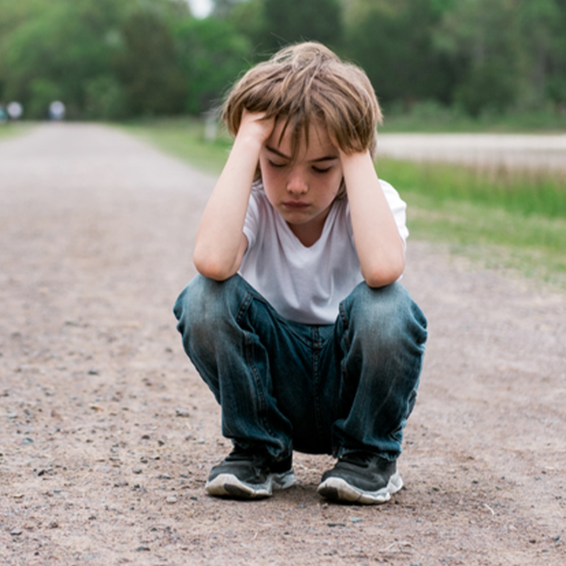Child crouching on a gravel path holding their head, appearing upset, outdoors with blurred trees in the background