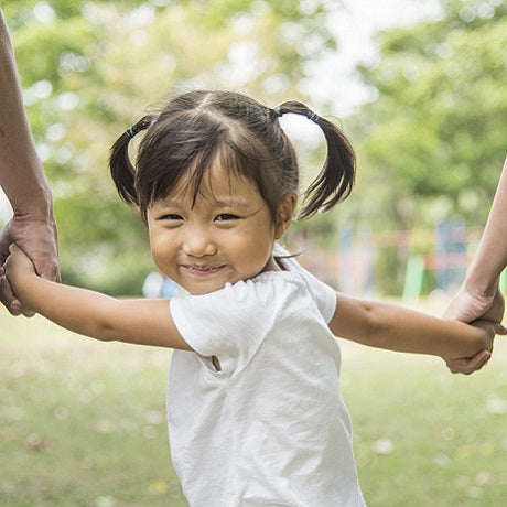 Smiling young girl with pigtails holding hands with two adults as they swing her between them in a sunlit park