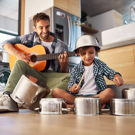 Father playing guitar while young child drums on pots in a kitchen