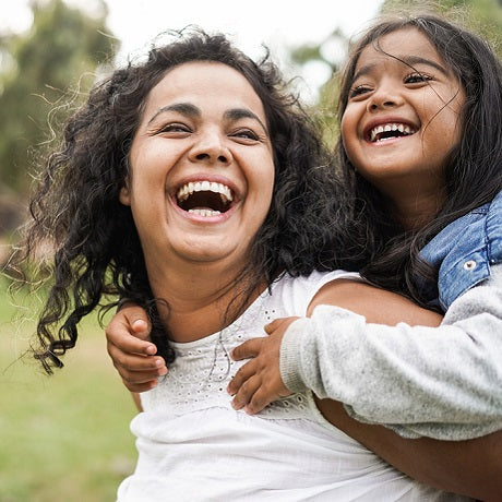Mother giving her daughter a piggyback ride while both laugh in a sunlit park