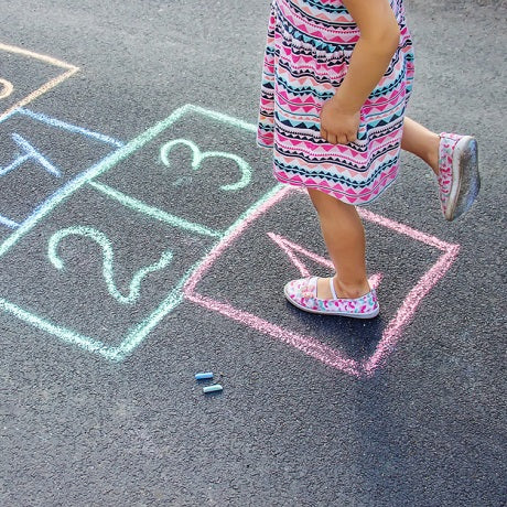 Child hopping in chalk hopscotch drawn on asphalt, wearing a pink patterned dress and floral slip-on shoes