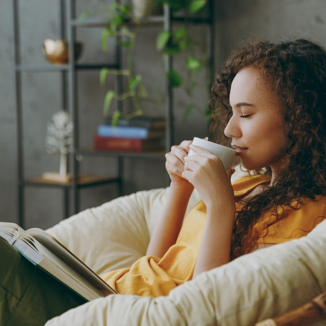Woman sitting in a cushioned armchair, sipping a mug with an open book on her lap, eyes closed and relaxed in a cozy room
