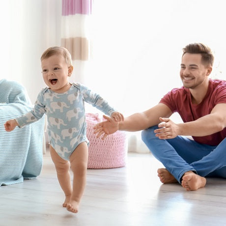 Toddler taking first steps toward a smiling father sitting on the floor in a bright nursery