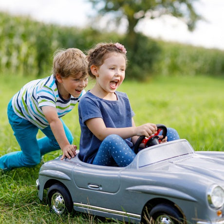Girl steering a toy ride-on car while a boy pushes from behind across a sunny grassy field, both laughing