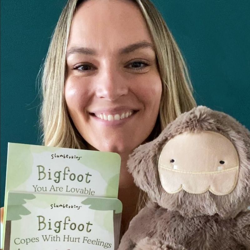 Woman holding a Bigfoot plush and two board books titled You Are Lovable and Copes With Hurt Feelings against a teal wall