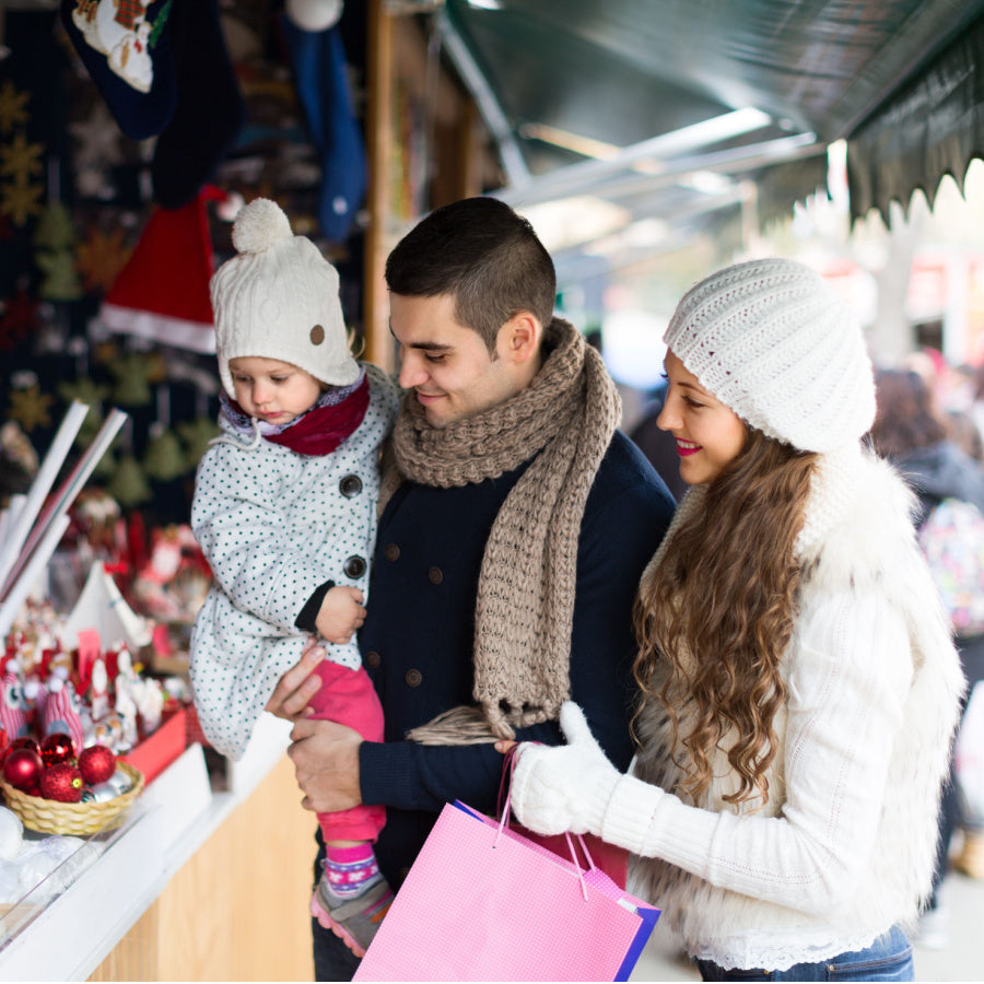 Parents with toddler browsing a festive holiday market stall in winter coats and hats, mother holding a pink shopping bag