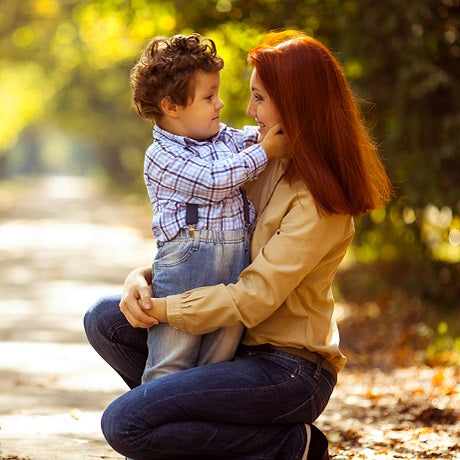 Mother kneeling on a sunlit park path listening attentively to a young child she holds