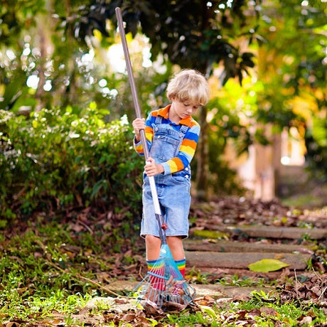 Child raking leaves on a garden path in denim overalls and a rainbow-striped sweater, sunny autumn yard