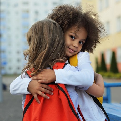 Two children hugging outdoors on a sidewalk, one wearing a red backpack, showing comfort and connection