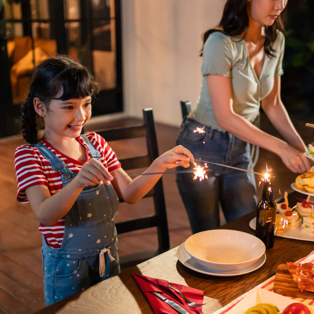 Girl holding lit sparklers at a family dinner table while an adult arranges food in the background