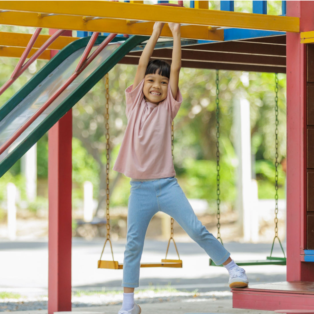 Child smiling while hanging from a colorful playground structure with swings and trees in the background