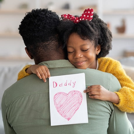 10 Tips to Teaching Kids About Mindful Gifting, girl hugging her father holding a card reading Daddy with a heart at home