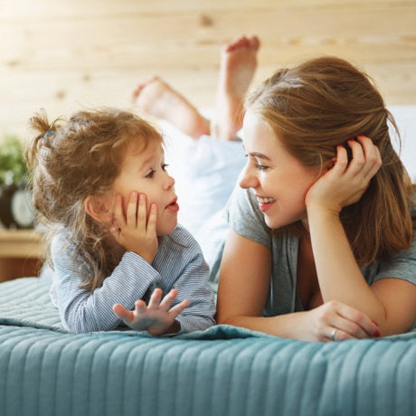 Woman and young child lying on a bed, facing each other and talking and smiling in a sunlit bedroom