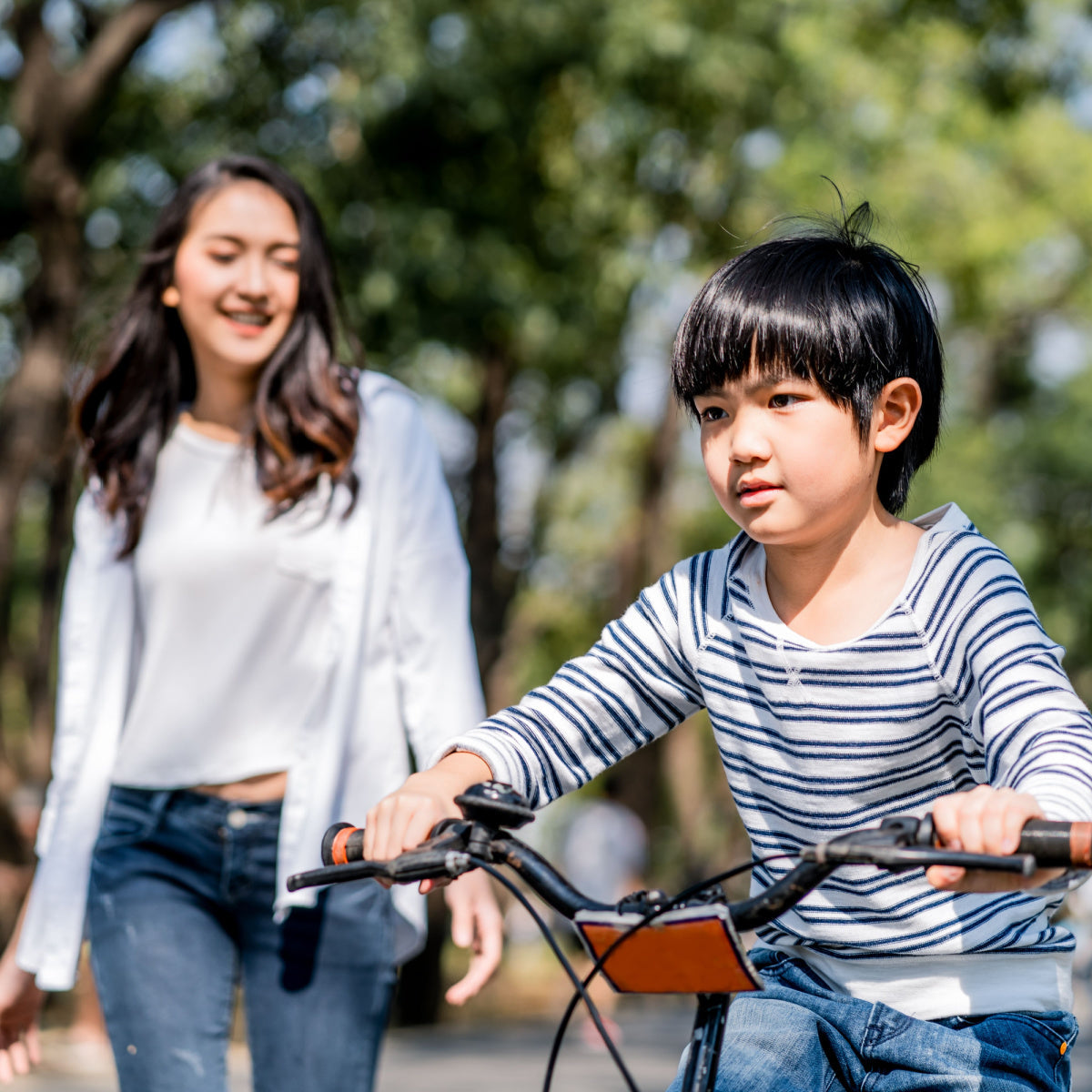 Child riding a bicycle in a sunny park while an adult caregiver walks behind offering encouragement