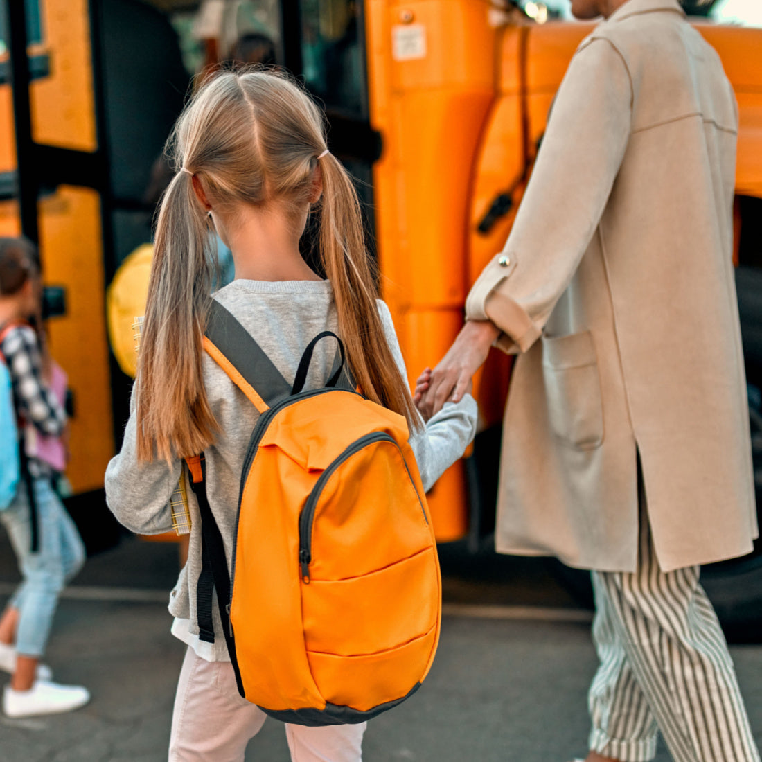Young girl with orange backpack holding an adult’s hand while boarding a yellow school bus
