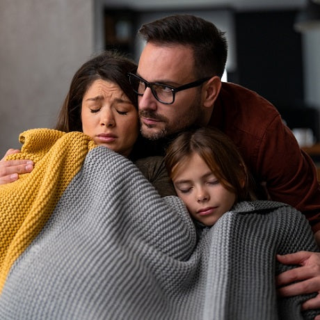 Parents comforting a child wrapped in gray and yellow blankets on a couch, appearing worried and supportive