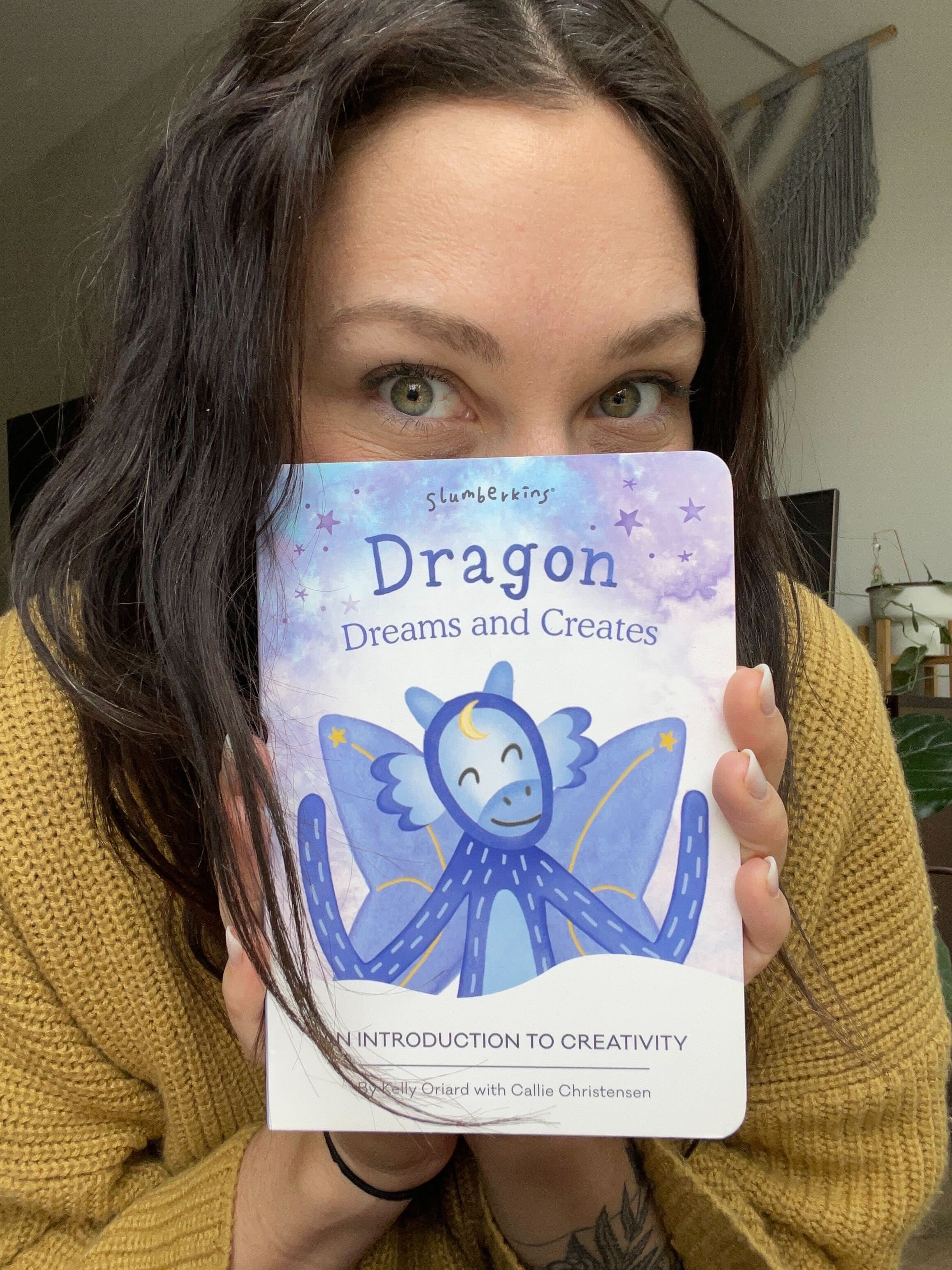 Woman peeking over a children’s board book titled ‘Dragon Dreams and Creates’ in a home setting