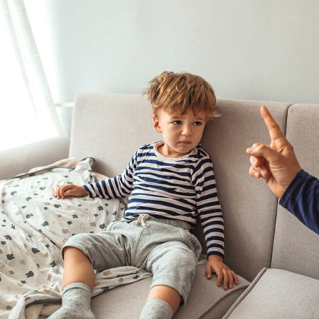 Young boy sitting on a couch looking upset as an adult points a finger in a scolding gesture