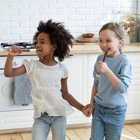 Two children sing into kitchen utensils while holding hands in a bright kitchen