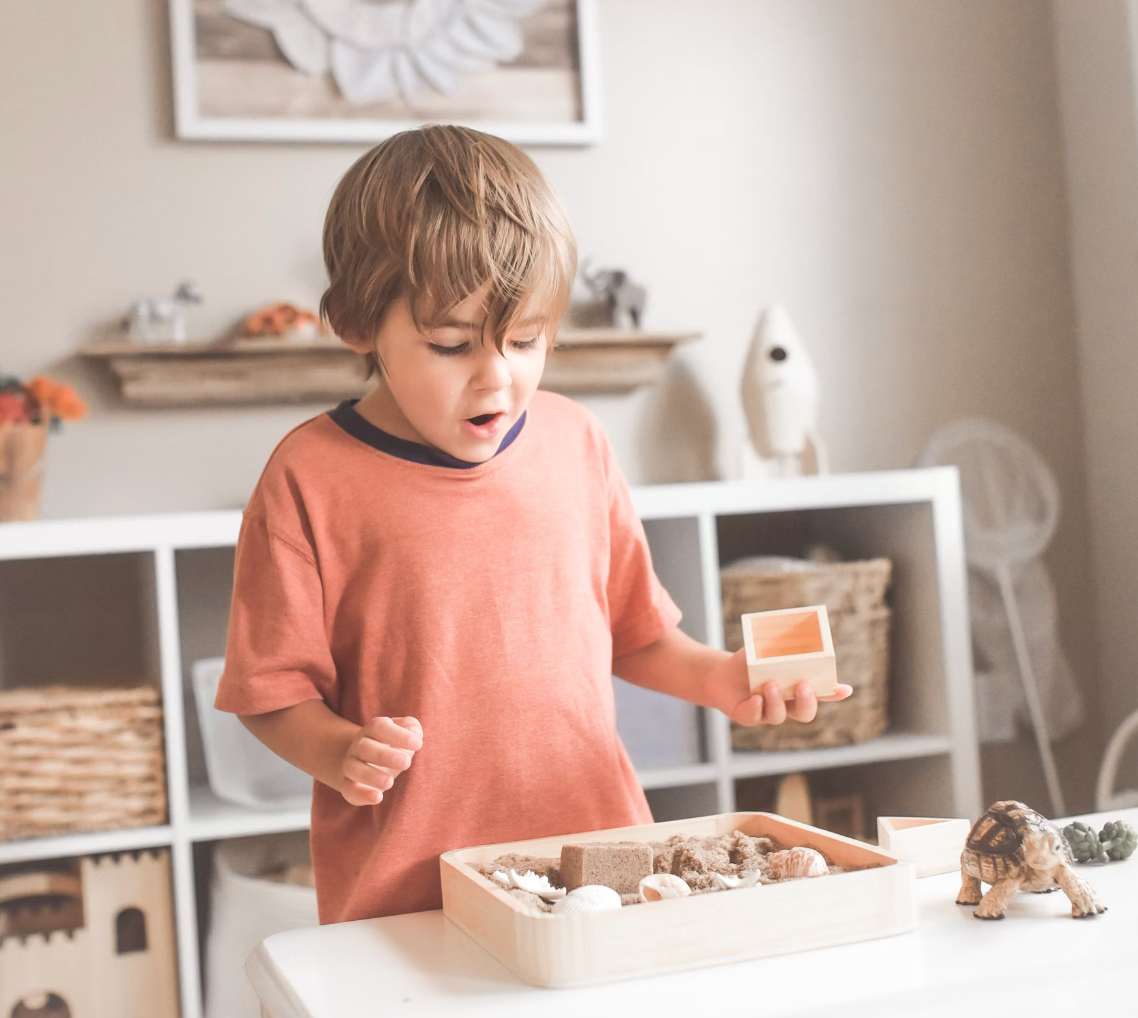 Young boy in a coral shirt holding a wooden block while exploring a sensory tray with sand and shells in a playroom