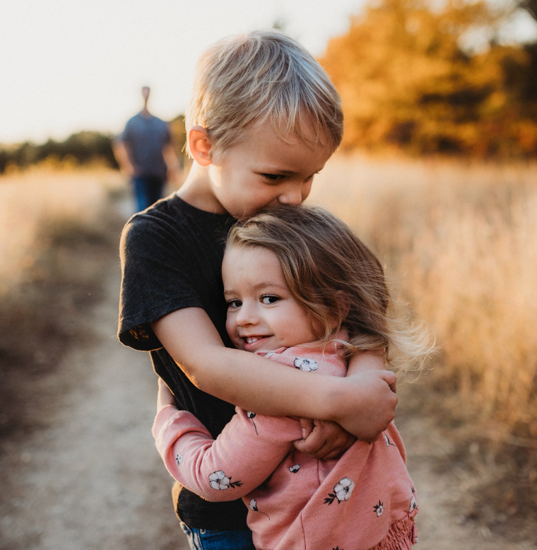 Two young siblings hugging on a dirt path at sunset, older child kissing the younger child’s head