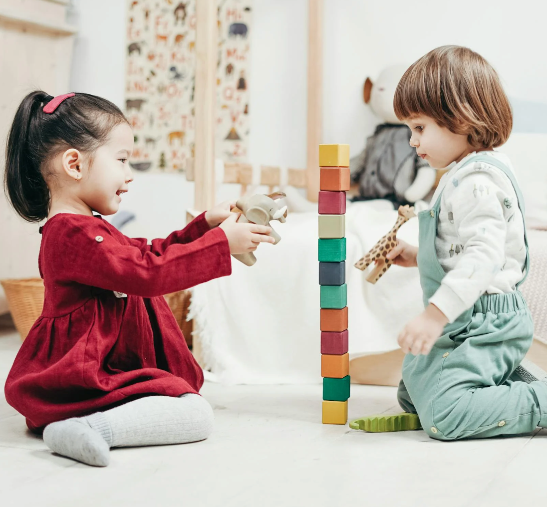 Two young children sitting on the floor playing and building a tall tower of colorful wooden blocks in a cozy bedroom play area