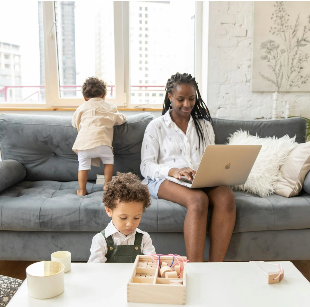 Mother working on a laptop on a living room couch while two young children play nearby at a coffee table