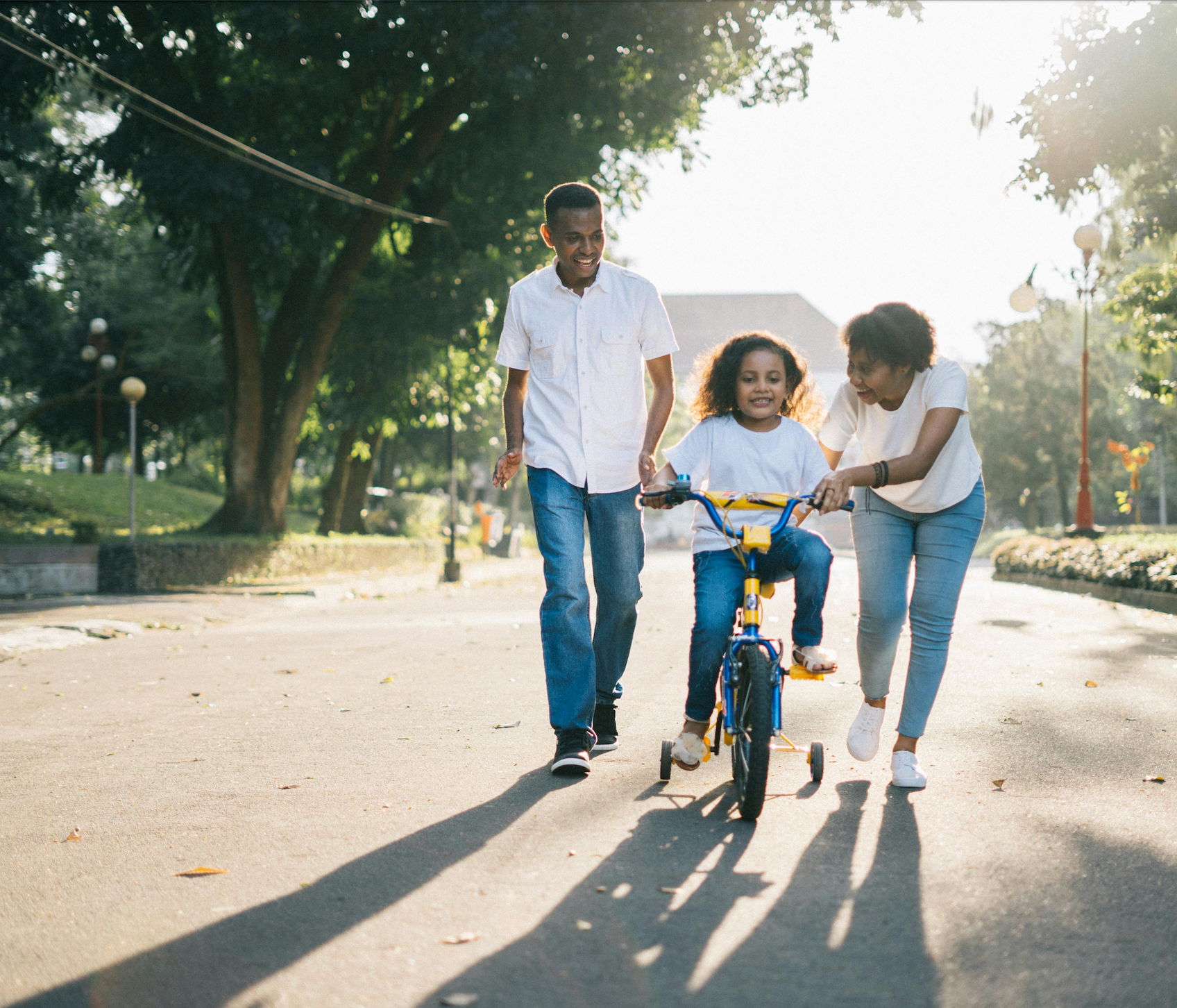 Child riding a bike with training wheels while parents run alongside and encourage them in a sunlit park