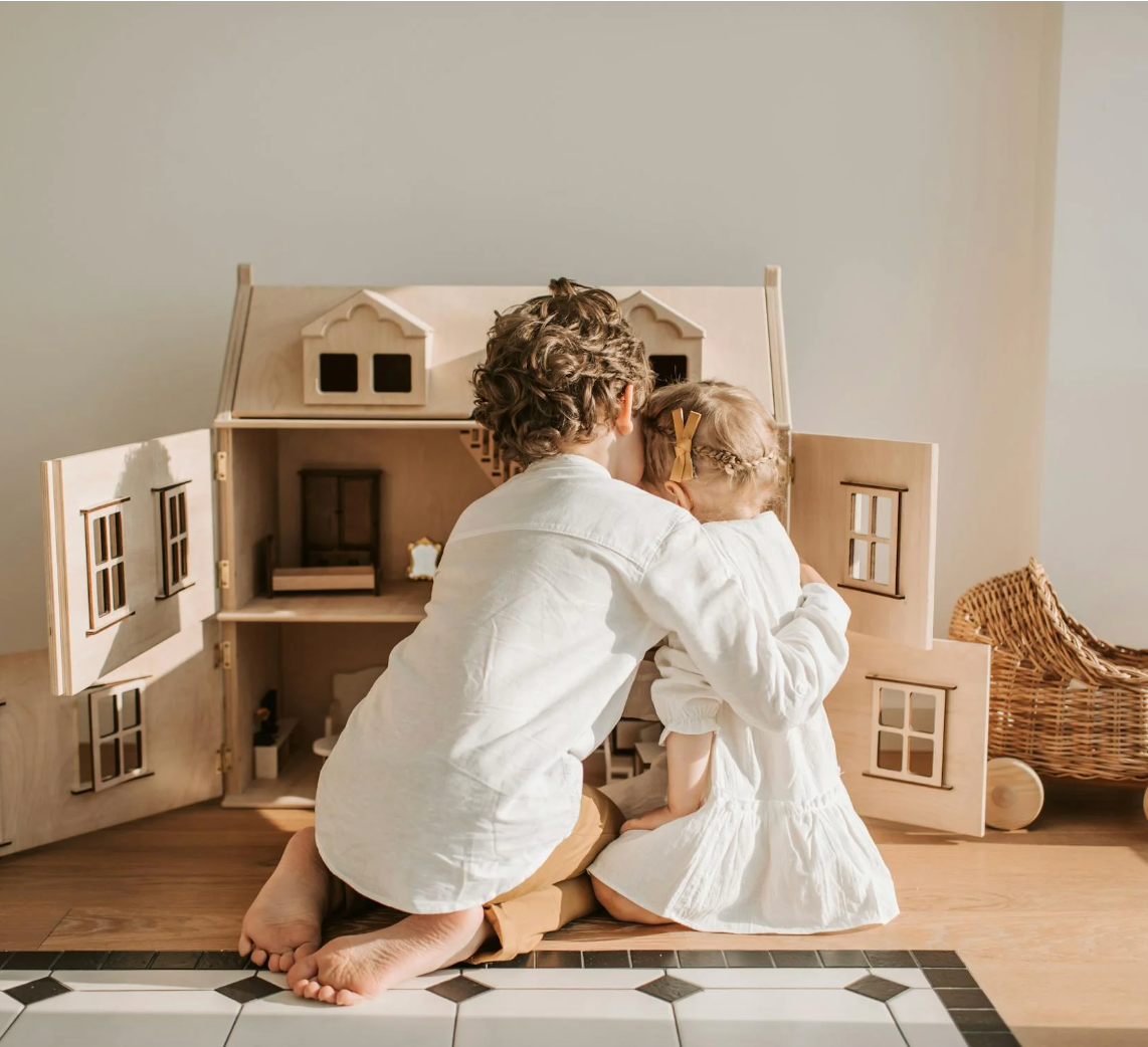 Two young children hugging while playing with a wooden dollhouse in a sunlit playroom