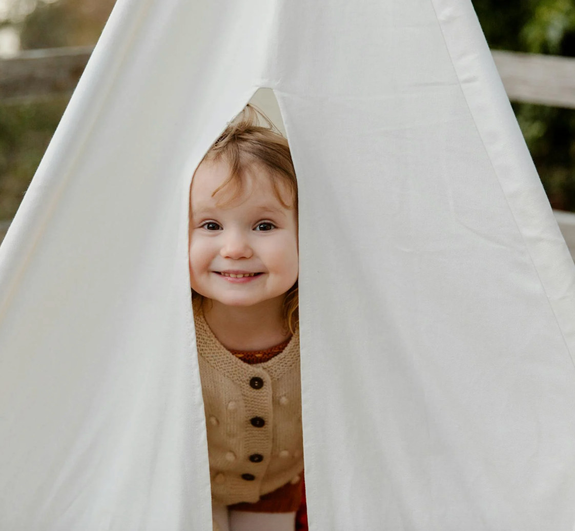 Toddler smiling and peeking through the opening of a white canvas play teepee outdoors