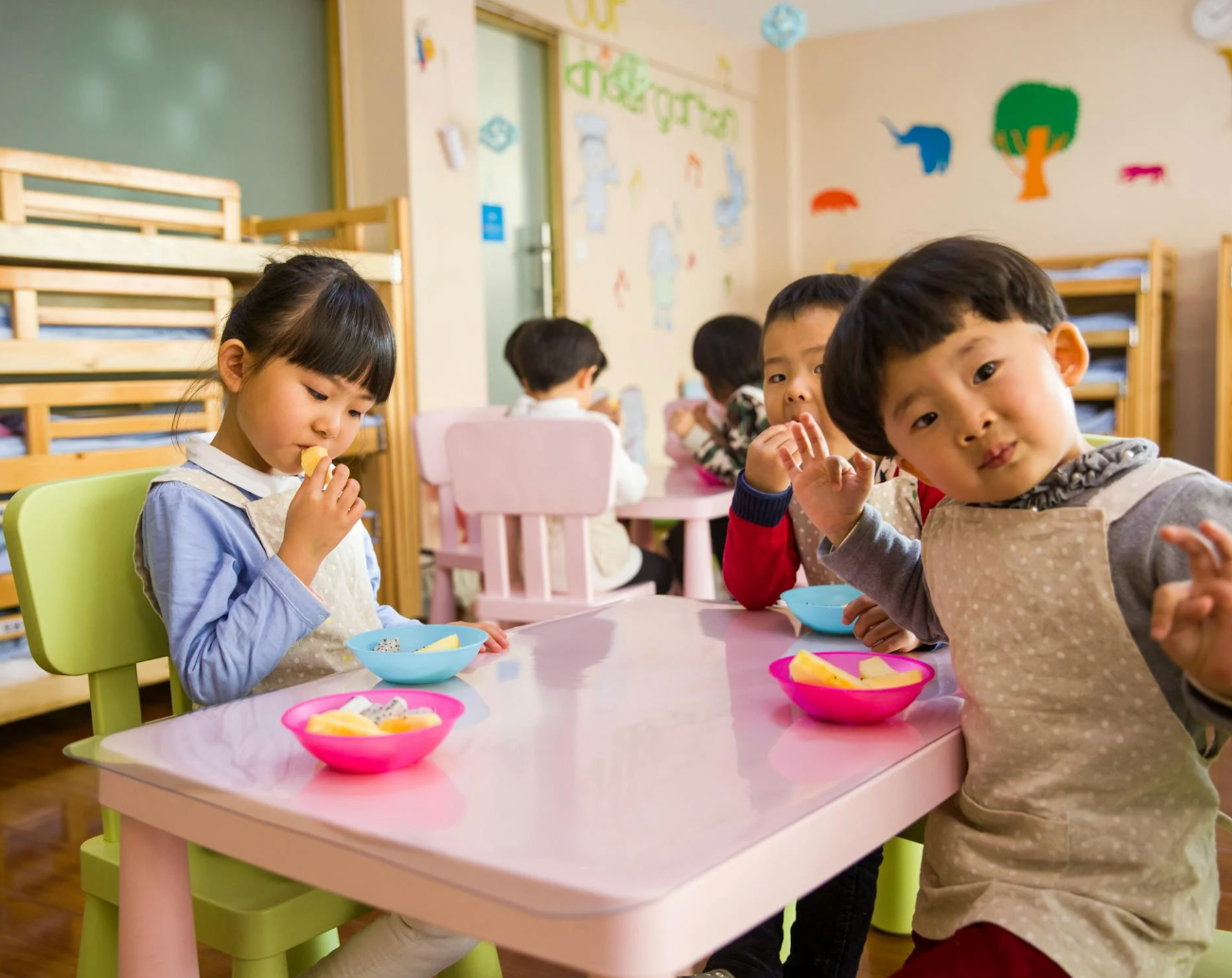 Preschool children eating snacks at a pink table in a classroom with colorful wall decorations, two children looking and waving at the camera