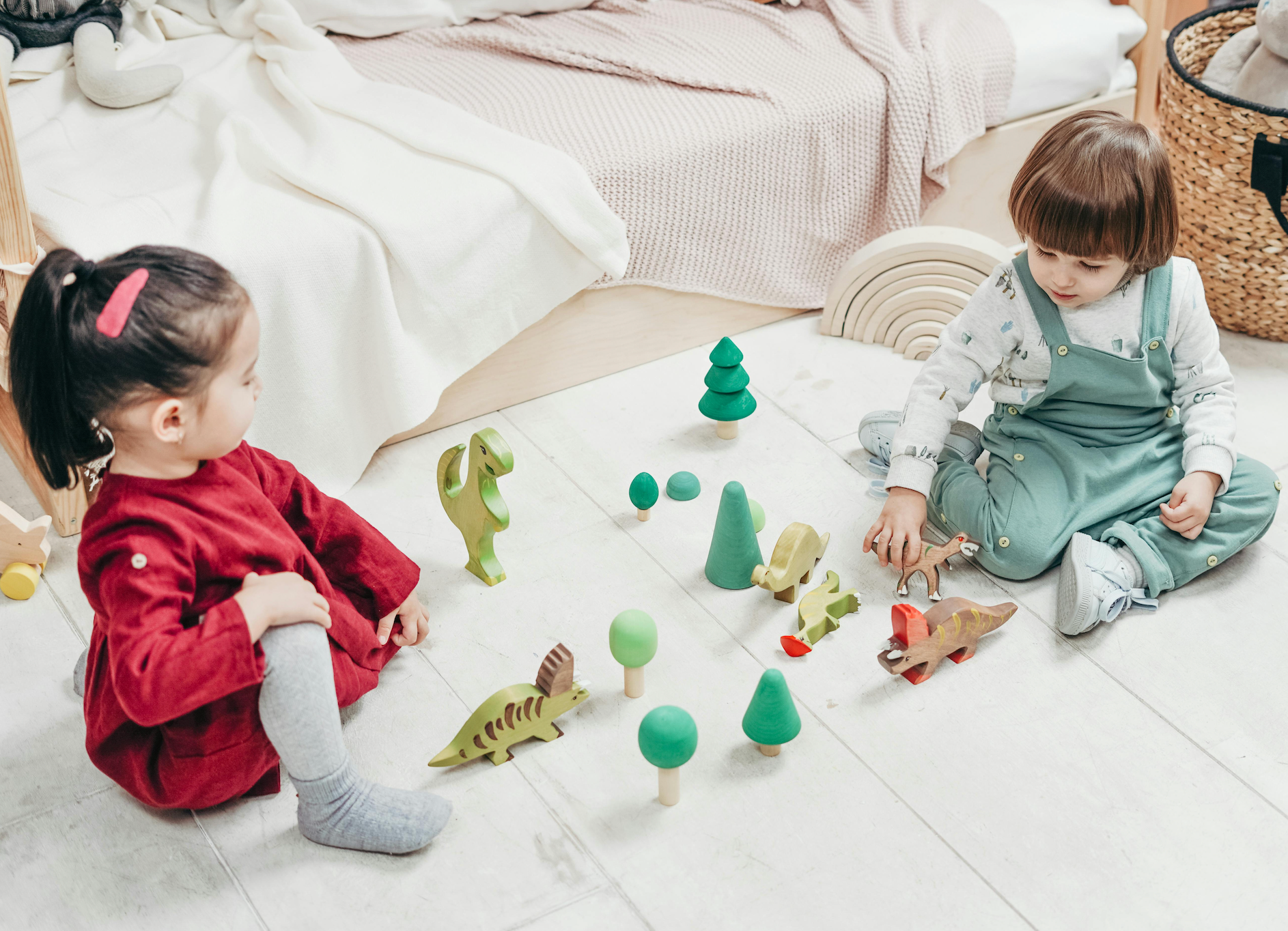 Two toddlers playing with wooden animal and tree toys on a light bedroom floor beside a bed