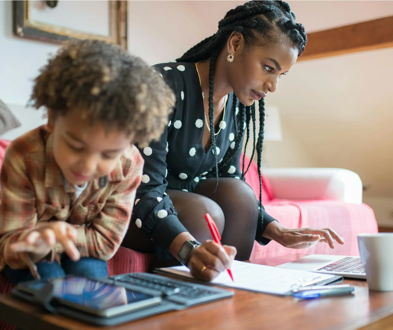 Mother working on a laptop and taking notes while her young child plays with a tablet on the living room couch