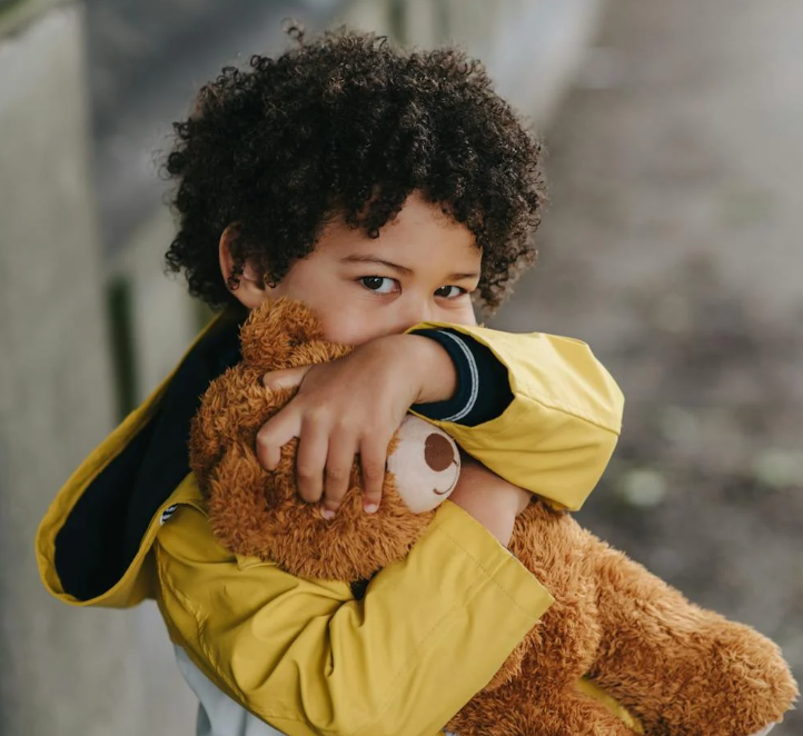 Young child hugging a brown stuffed bear outdoors, wearing a yellow raincoat and looking at the camera
