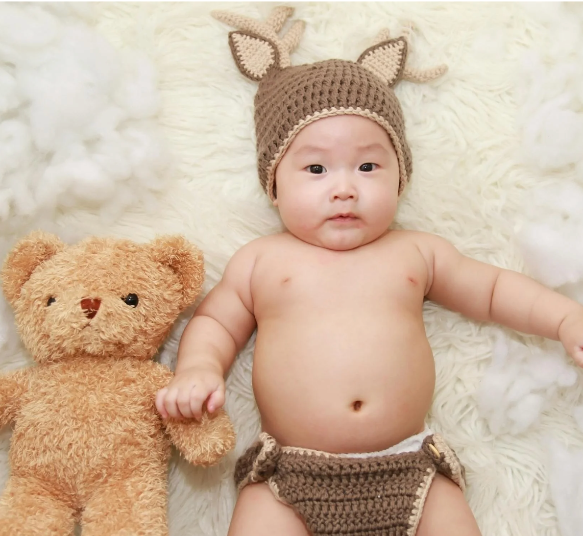 Baby lying on cream faux-fur rug holding a tan teddy bear, wearing a brown crocheted deer hat and matching diaper cover