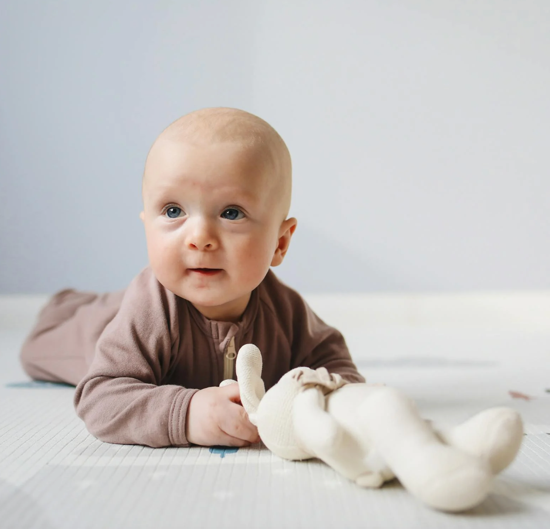 Baby lying on their tummy holding a cream knit stuffed animal on a play mat in a softly lit nursery