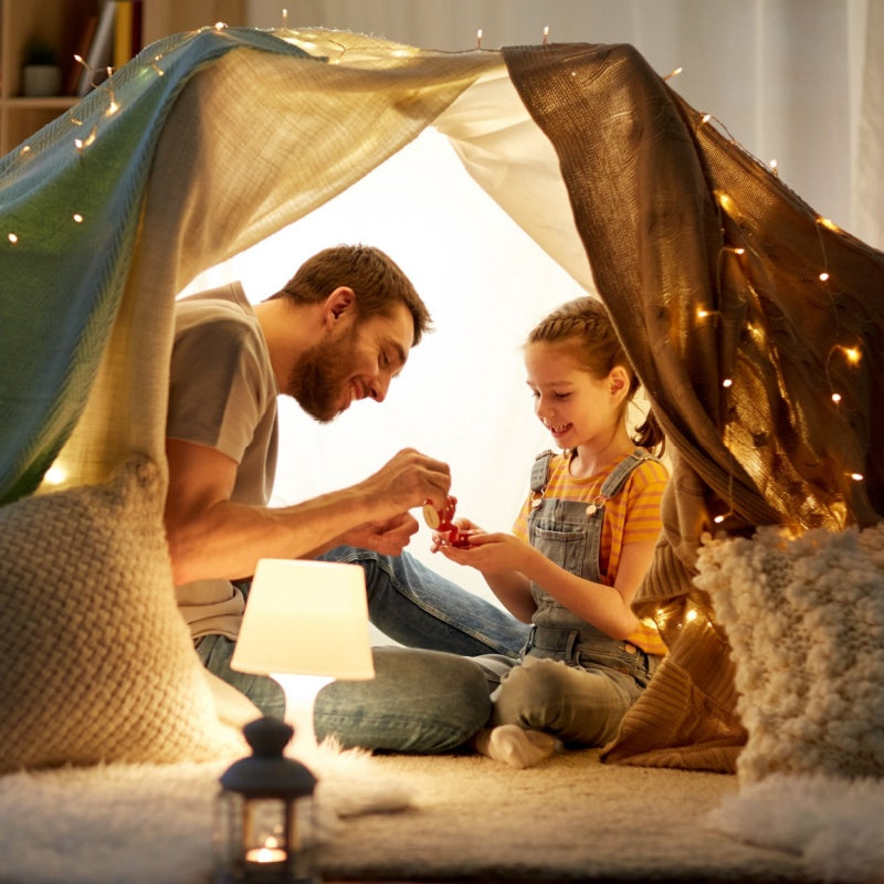 Father and young daughter playing inside a cozy blanket fort lit by string lights, sharing a small toy