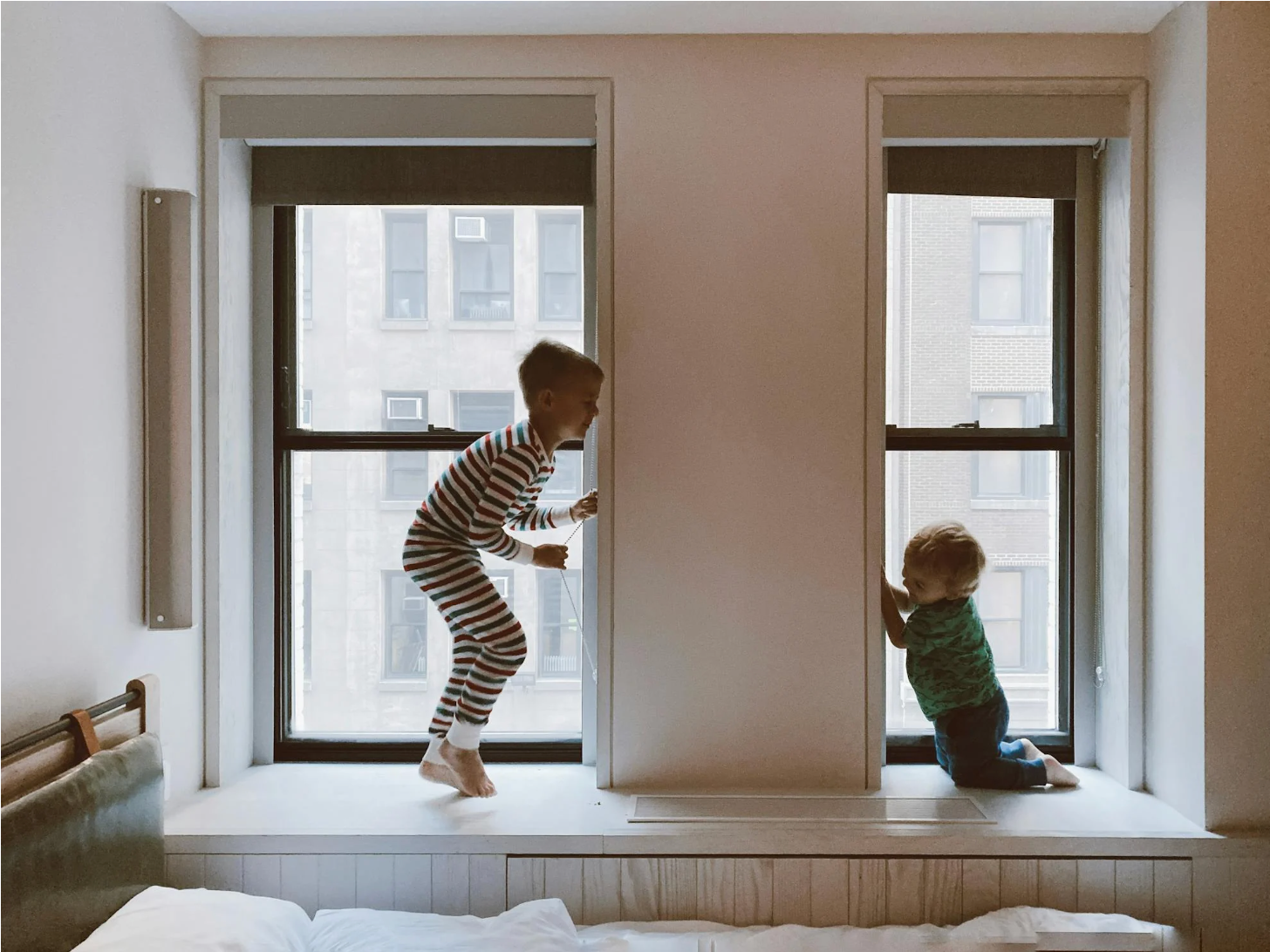 Two children playing on a window seat in a city apartment, an older boy jumping in striped pajamas while a toddler kneels by the window