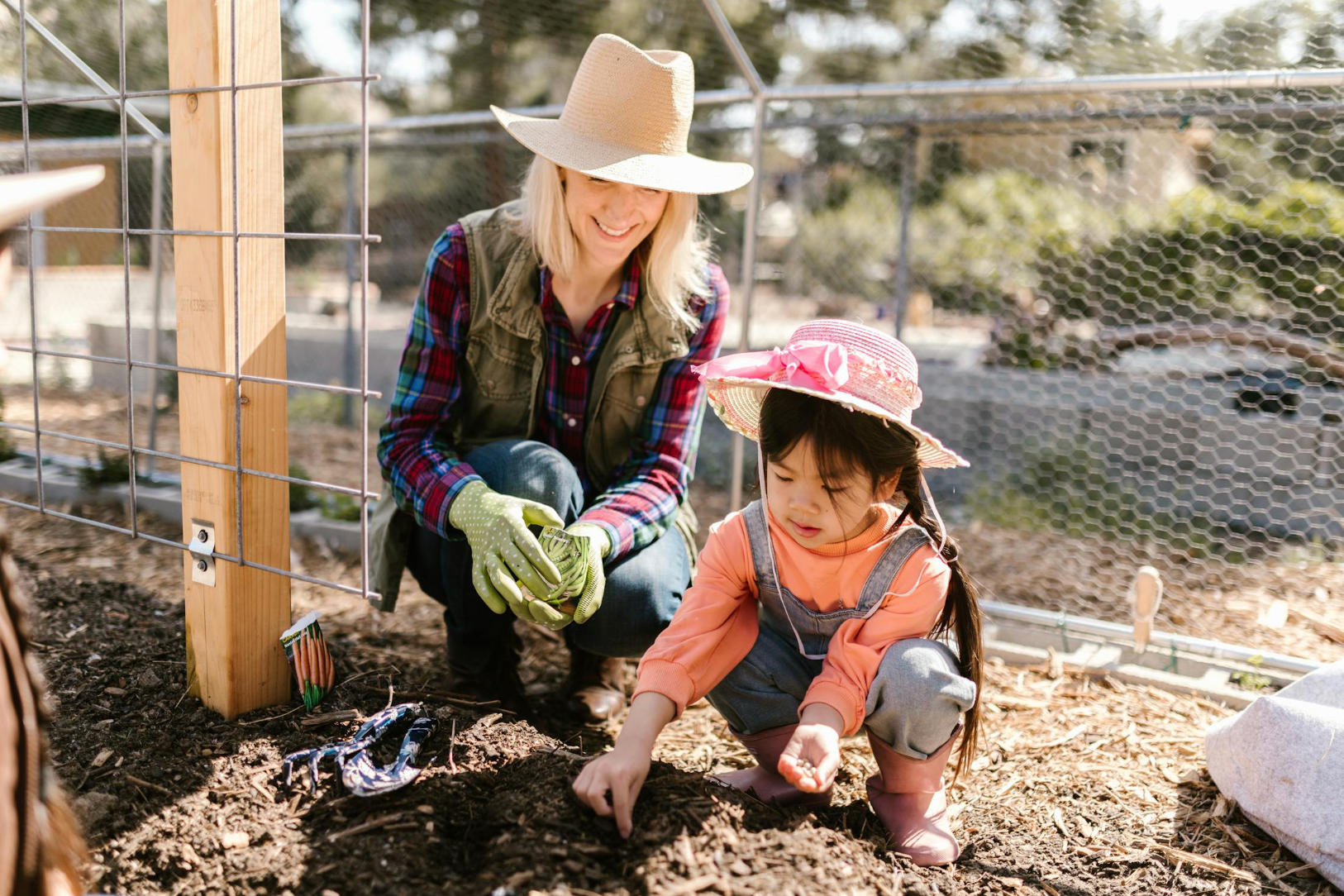 Woman kneeling with a young girl planting seeds in a fenced garden, child wearing a pink sun hat and boots