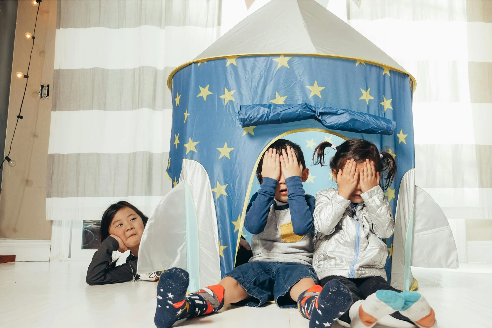 Two children covering their eyes while playing peekaboo in a blue star play tent as a sibling watches in a bright living room