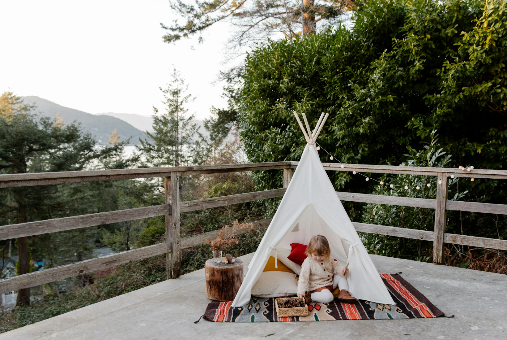 Child playing inside a small white teepee on a deck with string lights and a forested lake view in the background